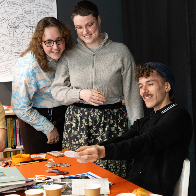 Three smiling participants at the badge station at QUEERCIRCLE in a photo by Christa Holka